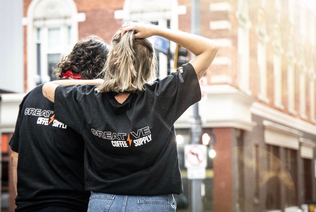 Two people wearing Creative Coffee Supply branded black t-shirts on an urban street