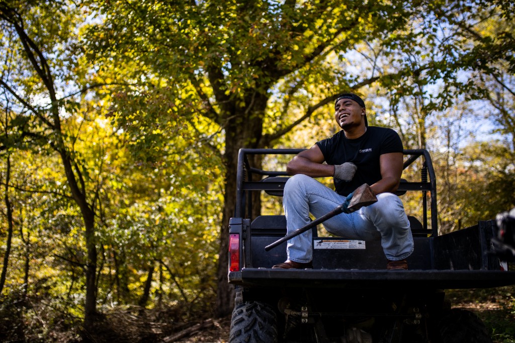 Tony Evans Jr. smiling on UTV with splitting maul across lap in autumn woods