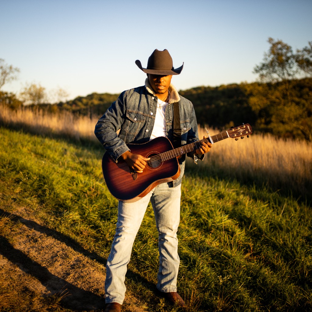 Full-length Tony Evans Jr. playing guitar in grassy field with warm sunlight