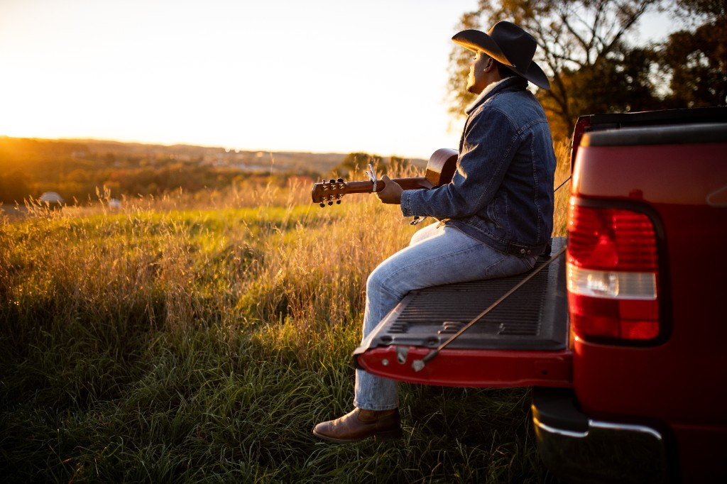 Side view of Tony Evans Jr. on truck tailgate with guitar at sunset