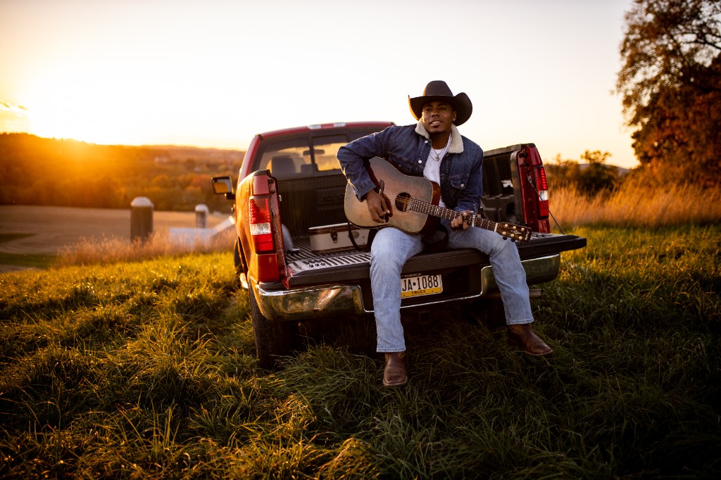 Tony Evans Jr. seated on truck tailgate playing acoustic guitar at sunset