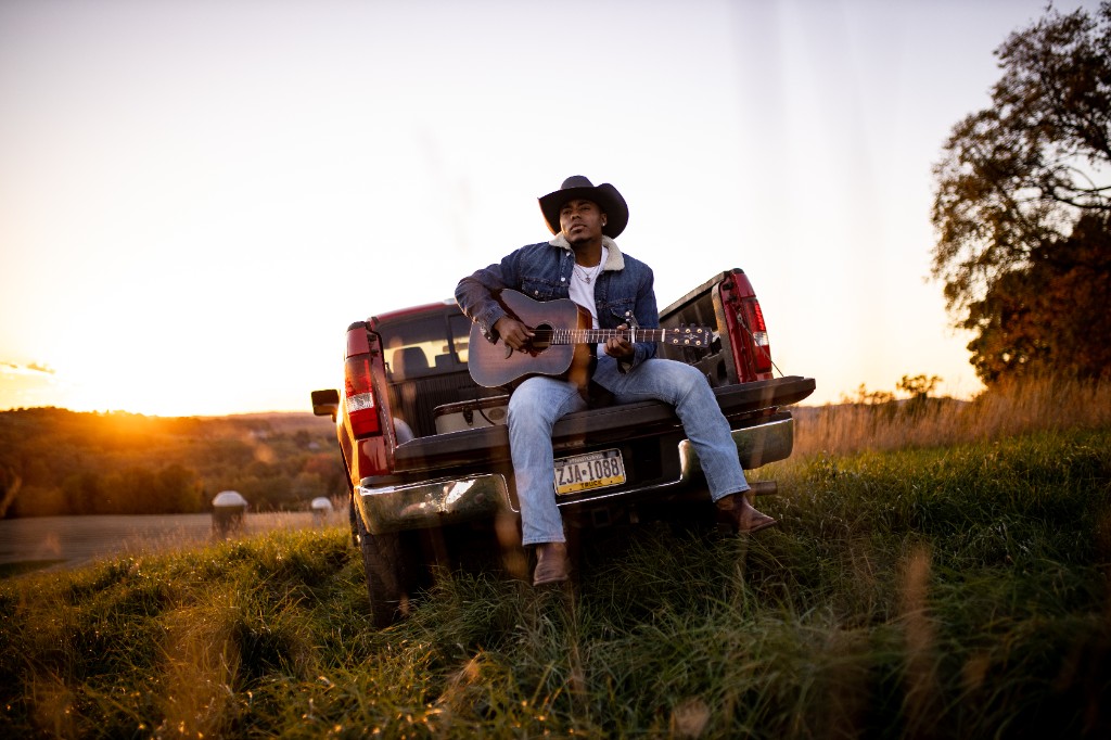 Tony Evans Jr. sitting on red pickup tailgate playing guitar at sunset in a field