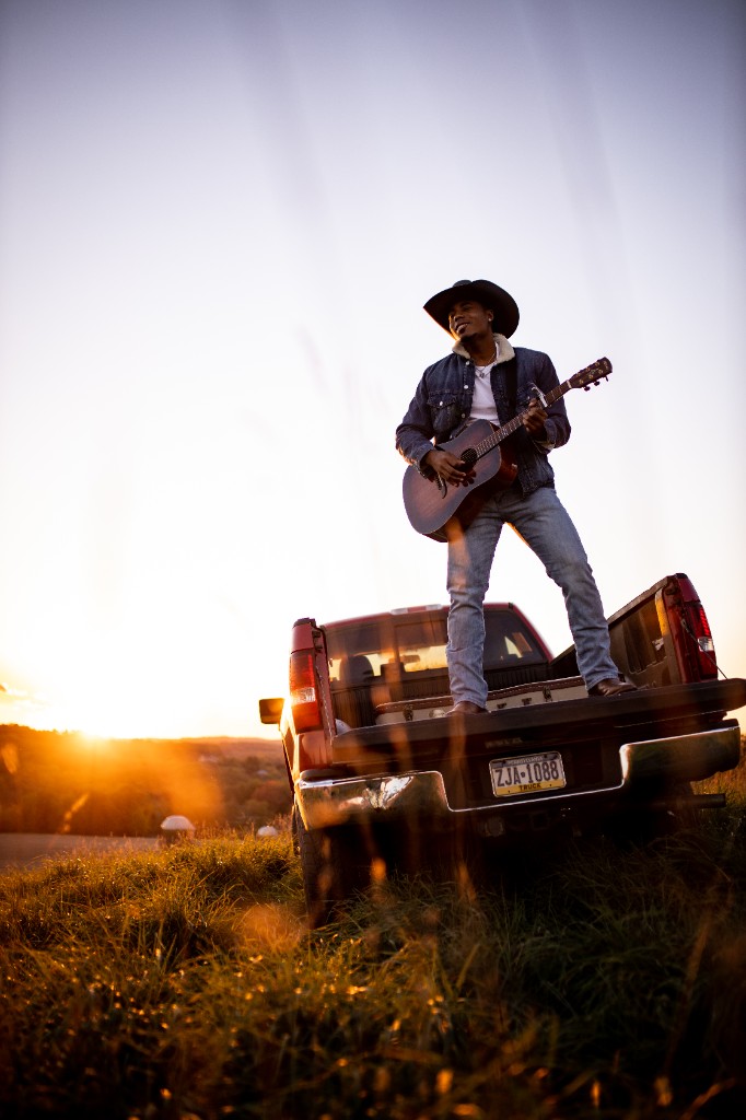 Low angle of Tony Evans Jr. standing in red truck bed playing guitar against bright sky