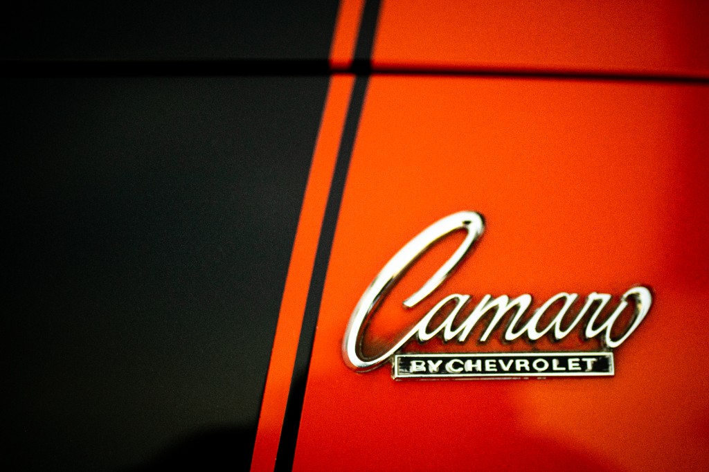 Chrome Camaro script emblem with BY CHEVROLET plate on orange paint beside black racing stripe